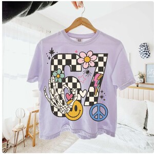 May include: A light purple t-shirt with a large graphic featuring the number "87" in a checkered pattern. The design includes a skeleton hand, flowers, a smiley face, a peace sign, and a lightning bolt. The shirt is hanging on a wooden hanger.