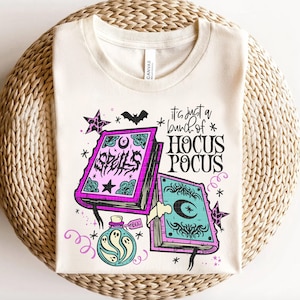 May include: Cream-coloured t-shirt with a Halloween-themed design. The graphic features two spell books, a potion bottle with ghosts, stars, and the phrase "It's just a bunch of Hocus Pocus." A bat and stars complete the design.