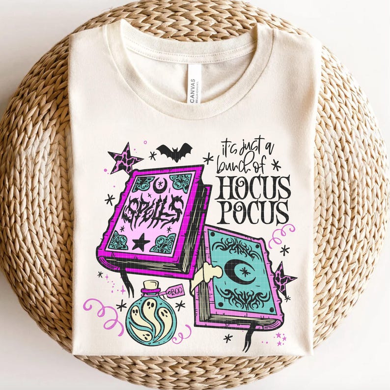 May include: Cream-coloured t-shirt with a Halloween-themed design. The graphic features two spell books, a potion bottle with ghosts, stars, and the phrase "It's just a bunch of Hocus Pocus." A bat and stars complete the design.