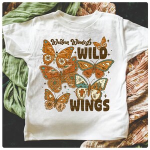 以下が含まれることがあります： オレンジと茶色の蝶のグラフィックと「Western Winds WILD WINGS」の文字が入った白いTシャツ。 蝶は詳細な模様があり、テキストの周りに配置されています。