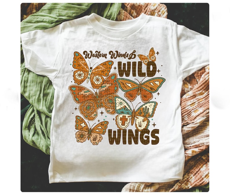 以下が含まれることがあります： オレンジと茶色の蝶のグラフィックと「Western Winds WILD WINGS」の文字が入った白いTシャツ。 蝶は詳細な模様があり、テキストの周りに配置されています。