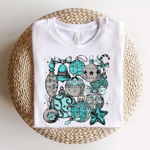 Peut inclure: T-shirt blanc avec un motif d'ornements de No&euml;l bleu sarcelle et gris. Les ornements sont d&eacute;cor&eacute;s d'&eacute;toiles, de volutes et d'autres d&eacute;tails festifs.