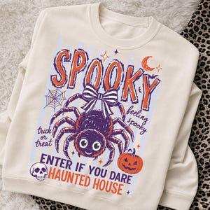 May include: A cream-colored sweatshirt featuring a graphic print with the text SPOOKY, trick or treat, feeling spooky, and ENTER IF YOU DARE HAUNTED HOUSE. The design includes a purple spider with a bow, a spiderweb, a skull, and an orange pumpkin.