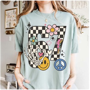 May include: A light blue t-shirt featuring a large graphic design. The design includes the number "87" in a black and white checkered pattern, a skeleton hand, a smiley face, flowers, a peace sign, and a lightning bolt.