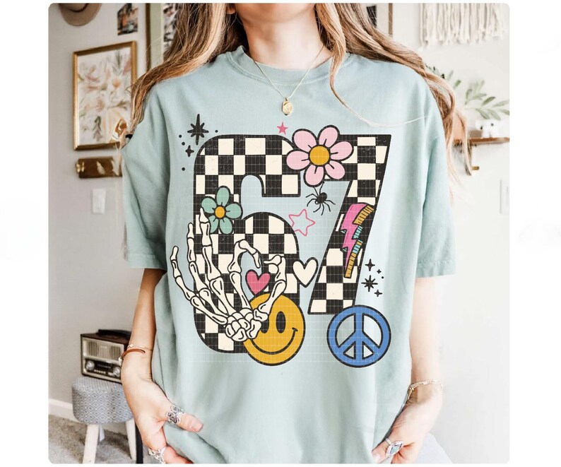 May include: A light blue t-shirt featuring a large graphic design. The design includes the number "87" in a black and white checkered pattern, a skeleton hand, a smiley face, flowers, a peace sign, and a lightning bolt.