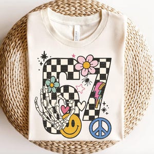 May include: Cream-colored t-shirt with a retro-style graphic featuring the number 67 in a black and white checkered pattern. The design includes a skeleton hand, smiley face, flowers, a peace sign, and a lightning bolt.