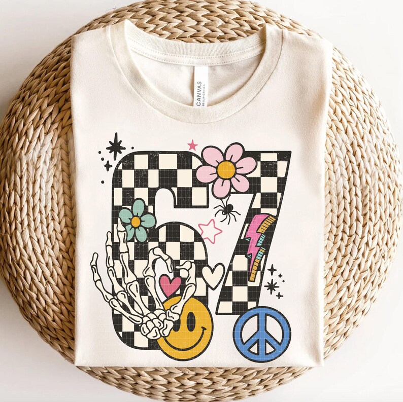 May include: Cream-colored t-shirt with a retro-style graphic featuring the number 67 in a black and white checkered pattern. The design includes a skeleton hand, smiley face, flowers, a peace sign, and a lightning bolt.