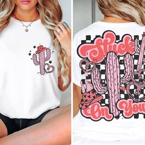 May include: White t-shirt with a pink cactus wearing a cowboy hat and the text "Stuck On You" on the back. The back design features a checkered pattern.