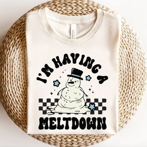 Pu&ograve; includere: Una maglietta bianca con una grafica nera di un pupazzo di neve che si scioglie con il testo "I'm having a meltdown".