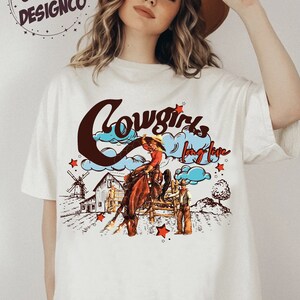 May include: Off-white t-shirt with a graphic of a cowboy riding a horse, a windmill, and the words "Cowgirls long live". The design is in brown, red, and blue tones. The text "GIZGIZ DESIGNCO" is in a circle at the top left.