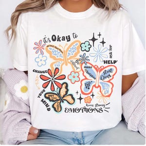 May include: White t-shirt with a colorful graphic design featuring butterflies, flowers, and the text "it's Okay to CHANGE your MIND, make Mistakes, be different, ask for 'HELP', Start Over, have strong EMOTIONS".