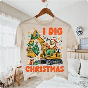 May include: Cream-colored t-shirt featuring a yellow excavator with a Christmas tree in its bucket and Santa Claus in the driver's seat. The text "I DIG CHRISTMAS" is printed in orange.