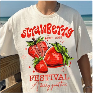 May include: White t-shirt with a red and green strawberry graphic and the text "Strawberry Festival" and "A really good time" in red.
