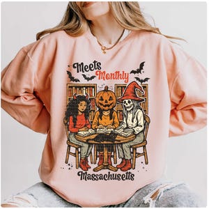 Może przedstawiać: Jasnor&oacute;żowa bluza z grafiką o tematyce Halloween. Grafika przedstawia trzy postacie wok&oacute;ł stołu, w tym diabła, dynię i szkielet, z napisem "Meets Monthly Massachusetts". W projekcie uwzględniono r&oacute;wnież nietoperze.