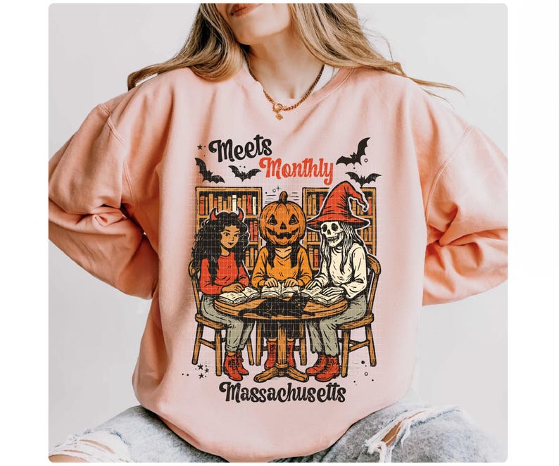 Może przedstawiać: Jasnor&oacute;żowa bluza z grafiką o tematyce Halloween. Grafika przedstawia trzy postacie wok&oacute;ł stołu, w tym diabła, dynię i szkielet, z napisem "Meets Monthly Massachusetts". W projekcie uwzględniono r&oacute;wnież nietoperze.