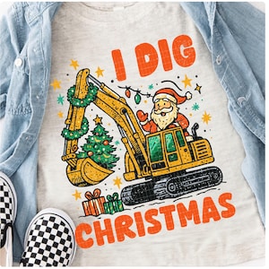 May include: A light gray t-shirt with the text "I DIG CHRISTMAS" in orange. The graphic features a yellow excavator with Santa Claus in the driver's seat, a Christmas tree, and wrapped gifts. The excavator has a green wreath and string lights.