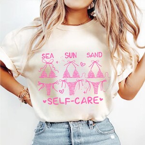 May include: Cream-colored t-shirt with a pink graphic featuring bikini designs and the words "SEA SUN SAND SELF-CARE." The design includes hearts and stars. The shirt is paired with blue jeans.