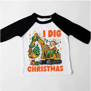 May include: White and black raglan-style shirt with a colorful graphic. The graphic features a yellow excavator with Santa Claus in the driver's seat, a Christmas tree, and the text "I DIG CHRISTMAS" in orange.