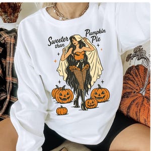 Peut inclure: Sweat-shirt blanc &agrave; manches longues avec un motif d'Halloween. Le motif repr&eacute;sente une femme en costume, des citrouilles et le texte "Sweeter than Pumpkin Pie". La femme porte un corset, des bas r&eacute;sille et une cape.