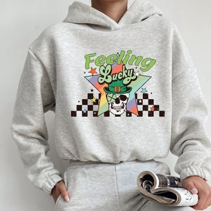 K&ouml;nnte beinhalten: Ein grauer Hoodie mit einem grafischen Druck eines Sch&auml;dels, der einen gr&uuml;nen Koboldhut tr&auml;gt, und dem Text "Feeling Lucky" in einem Regenbogen-Stern-Design.