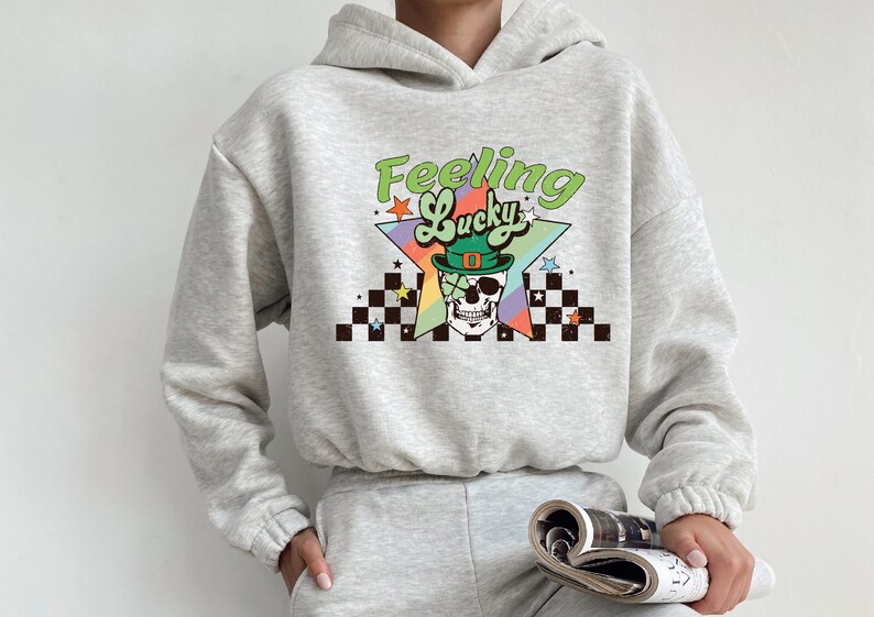K&ouml;nnte beinhalten: Ein grauer Hoodie mit einem grafischen Druck eines Sch&auml;dels, der einen gr&uuml;nen Koboldhut tr&auml;gt, und dem Text "Feeling Lucky" in einem Regenbogen-Stern-Design.