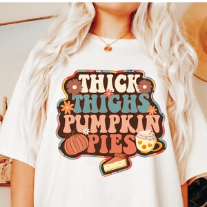 May include: White t-shirt featuring a retro-style graphic that reads "Thick Thighs Pumpkin Pies". The graphic includes a pumpkin, a cup of coffee, and a slice of pie.