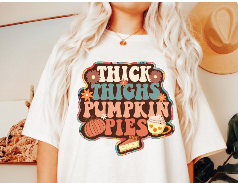 May include: White t-shirt featuring a retro-style graphic that reads "Thick Thighs Pumpkin Pies". The graphic includes a pumpkin, a cup of coffee, and a slice of pie.