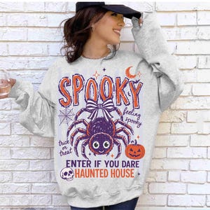 May include: A light gray crewneck sweatshirt featuring a graphic print with the text SPOOKY, feeling spooky, trick or treat, ENTER IF YOU DARE, and HAUNTED HOUSE. The design includes a purple spider with a bow, a spiderweb, a crescent moon, and a pumpkin.