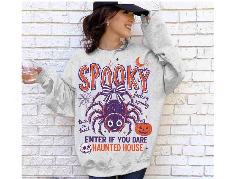 May include: A light gray crewneck sweatshirt featuring a graphic print with the text SPOOKY, feeling spooky, trick or treat, ENTER IF YOU DARE, and HAUNTED HOUSE. The design includes a purple spider with a bow, a spiderweb, a crescent moon, and a pumpkin.