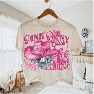 May include: Cream-colored t-shirt with a pink graphic. The graphic features a pink cowboy hat, a disco ball, and three pink horses. The text "PINK PONY CLUB" is above the hat. The shirt is hanging on a wooden hanger.