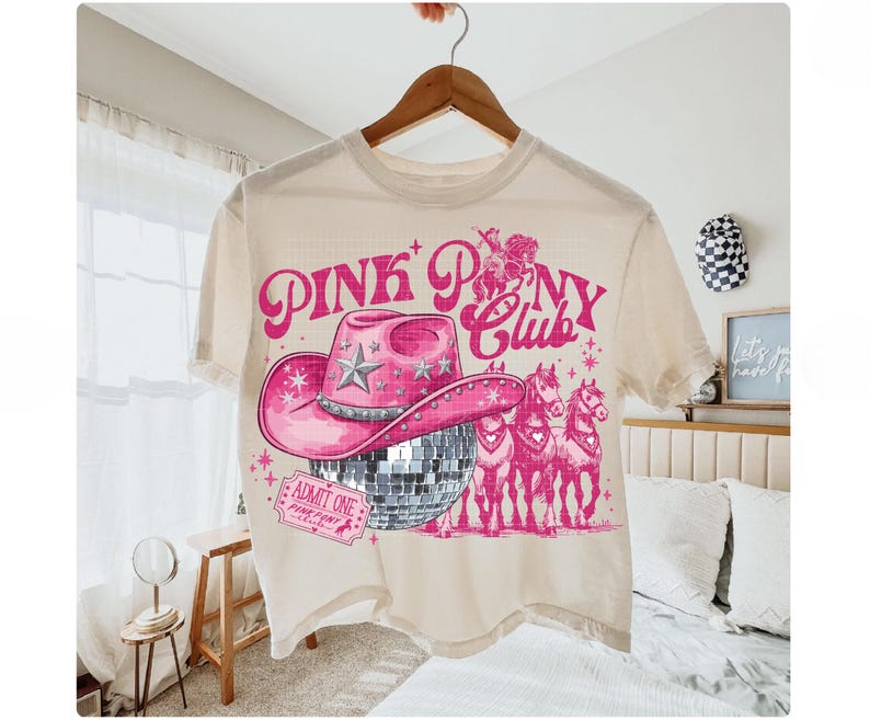 May include: Cream-colored t-shirt with a pink graphic. The graphic features a pink cowboy hat, a disco ball, and three pink horses. The text "PINK PONY CLUB" is above the hat. The shirt is hanging on a wooden hanger.