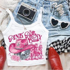 May include: White tank top with a pink graphic that reads "PINK PONY CLUB" featuring a pink cowboy hat, disco ball, and horses. The image also includes red cowboy boots, denim shorts, a camera, and heart-shaped sunglasses.