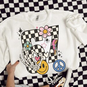 May include: White crewneck sweatshirt with a colorful graphic design. The design includes a checkered "7" with flowers, a skeleton hand, a smiley face, a peace sign, and a lightning bolt. The sweatshirt is on a black and white checkered surface.