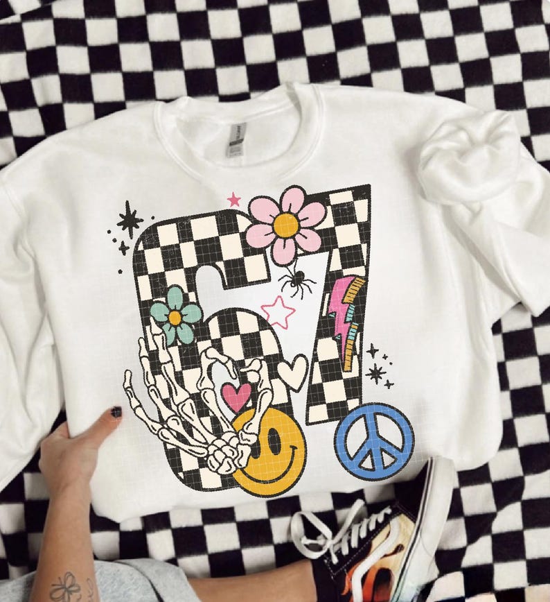 May include: White crewneck sweatshirt with a colorful graphic design. The design includes a checkered "7" with flowers, a skeleton hand, a smiley face, a peace sign, and a lightning bolt. The sweatshirt is on a black and white checkered surface.