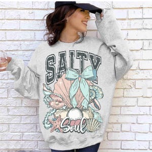 Puede incluir: Sudadera gris con un gr&aacute;fico de conchas marinas y un lazo, y el texto "Salty Soul".