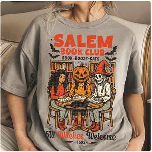 Może przedstawiać: Szara koszulka z grafiką o tematyce Halloween. Projekt zawiera napis "Salem Book Club Book-Booze-Bats" w kolorze pomarańczowym, z ilustracjami czarownic, postaci z dyniową głową i szkieletu czytającego książki. Zawiera r&oacute;wnież frazę "All Witches Welcome 1692".