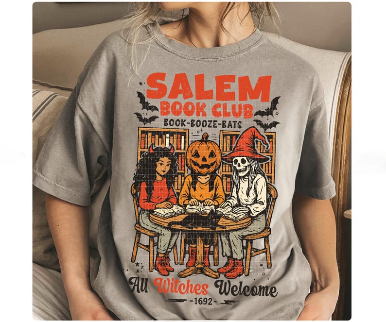 Może przedstawiać: Szara koszulka z grafiką o tematyce Halloween. Projekt zawiera napis "Salem Book Club Book-Booze-Bats" w kolorze pomarańczowym, z ilustracjami czarownic, postaci z dyniową głową i szkieletu czytającego książki. Zawiera r&oacute;wnież frazę "All Witches Welcome 1692".