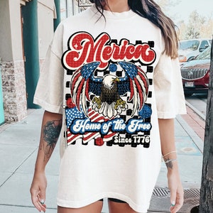 May include: White t-shirt with a red, white, and blue graphic design featuring an eagle, American flags, and the text "Merica Home of the Free Since 1776".