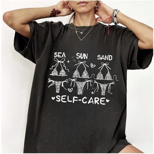 May include: Dark gray t-shirt with the words "SEA SUN SAND SELF-CARE" in white. The shirt features a graphic of three bikini tops and bottoms. The design includes hearts and stars.