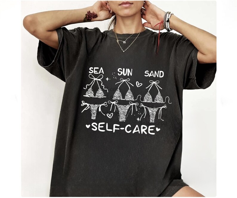 May include: Dark gray t-shirt with the words "SEA SUN SAND SELF-CARE" in white. The shirt features a graphic of three bikini tops and bottoms. The design includes hearts and stars.