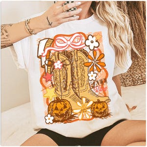 May include: White t-shirt featuring a graphic design with cowboy boots, pumpkins, and floral elements in shades of orange, brown, and white. The text "Fall Vibes" is also included in the design.