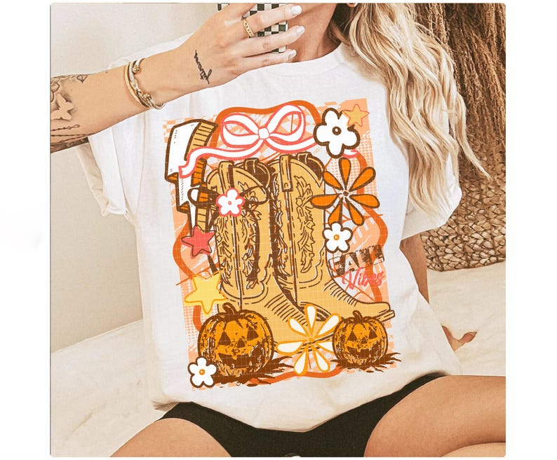 May include: White t-shirt featuring a graphic design with cowboy boots, pumpkins, and floral elements in shades of orange, brown, and white. The text "Fall Vibes" is also included in the design.