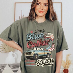 May include: Olive green t-shirt with a graphic design featuring a blue pickup truck, a baseball cap, and boots. The text reads "Living Blue Collar" and "Dirty Hands Clean Money".