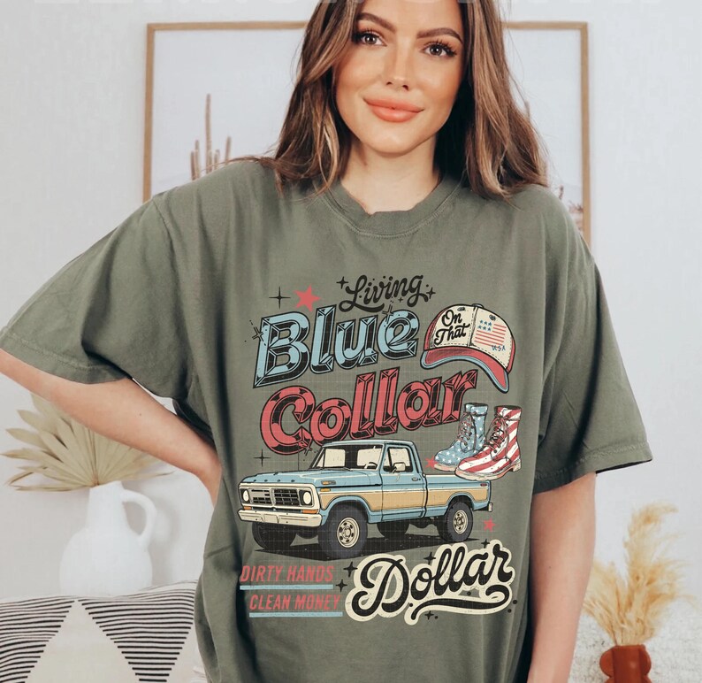 May include: Olive green t-shirt with a graphic design featuring a blue pickup truck, a baseball cap, and boots. The text reads "Living Blue Collar" and "Dirty Hands Clean Money".