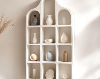 May include: A white, arched wall shelf with multiple square compartments, each holding a small decorative vase. The vases vary in shape, color, and texture, including speckled, smooth, and cylindrical designs. The shelf is mounted on a neutral-colored wall.