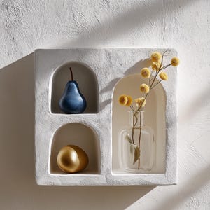 Può includere: Un ripiano a muro in gesso bianco con tre aperture ad arco. Il ripiano contiene una pera blu, un uovo dorato e un vaso di vetro trasparente con fiori gialli secchi.