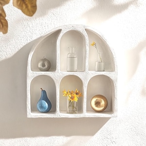 Peut inclure: Une étagère murale blanche en forme d'arche avec six compartiments contenant une sphère grise, une bouteille en verre transparent, un petit vase avec une fleur jaune, une poire bleue, un vase avec des fleurs jaunes et une sphère dorée.