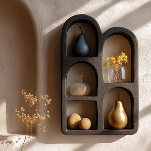 May include: A black ceramic wall shelf with six arched compartments. The shelf is decorated with a blue pear, a clear glass vase with a white textured vase, a bouquet of dried flowers in a clear glass vase, and two gold pears.