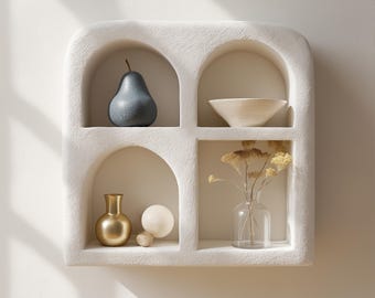 Peut inclure: Une étagère murale en plâtre blanc avec quatre niches en forme d'arche. La niche supérieure gauche contient une poire bleue, la niche supérieure droite contient un bol en céramique blanche, la niche inférieure gauche contient un vase doré et une sphère blanche, et la niche inférieure droite contient un vase en verre transparent avec des fleurs séchées.