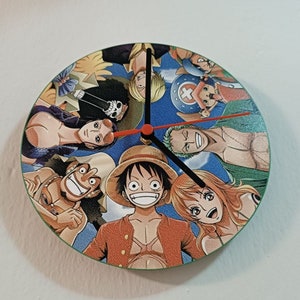May include: A round clock with a blue background featuring characters from the anime One Piece. The clock has black hands and a red second hand. The characters include Luffy, Zoro, Nami, Usopp, Sanji, Chopper, and Franky.